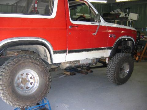 1982 Ford Bronco - A red and white two-door Ford Bronco SUV shown from the passenger side, lifted on a jack with oversized mud tires on silver wheels, inside…