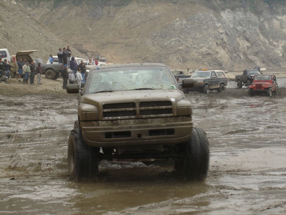 Muddy Dodge Ram pickup truck with large tires driving through a muddy area. Other trucks and people are visible in the background