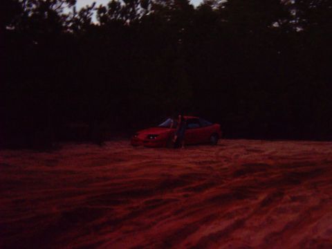 1993 Ford Bronco - A red coupe is parked in a sandy area with trees in the background, and a person is leaning against the driver's door.