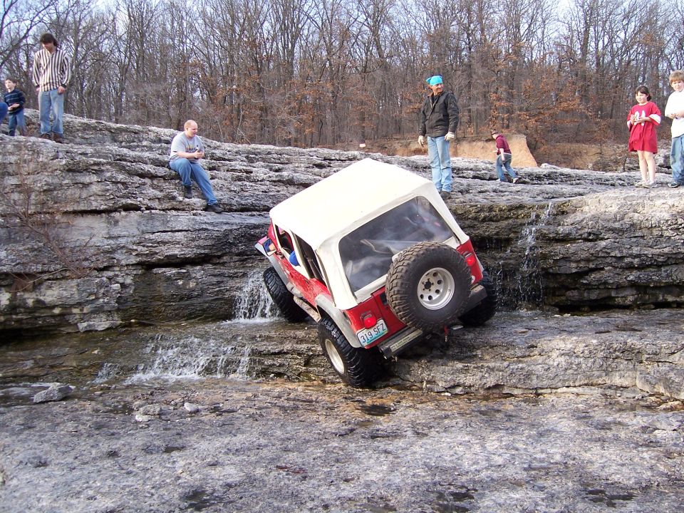 Red Jeep with white hardtop nose-down on wet rocky ledge as people watch.