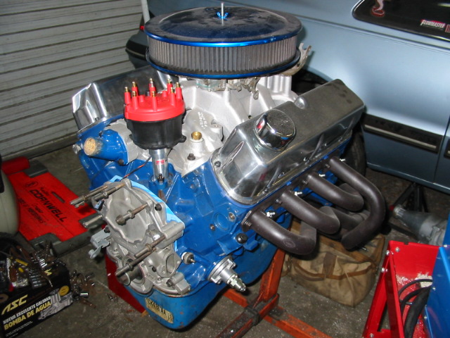 Blue V8 engine with chrome valve covers and air cleaner on an engine stand, next to a light blue car