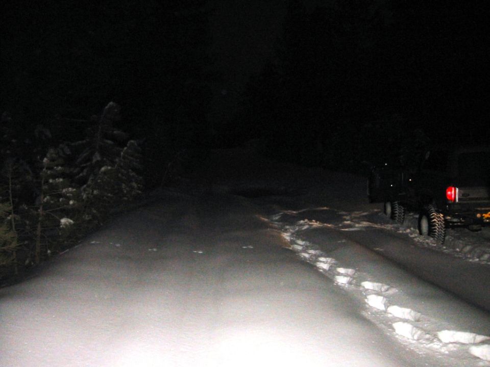 Dark pickup with large tires parked on a snowy road at night, footprints in the snow