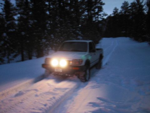 1999 Toyota Tacoma - A 1999 Toyota Tacoma navigates a snow-covered road as dusk settles, its headlights cutting through the dim light.