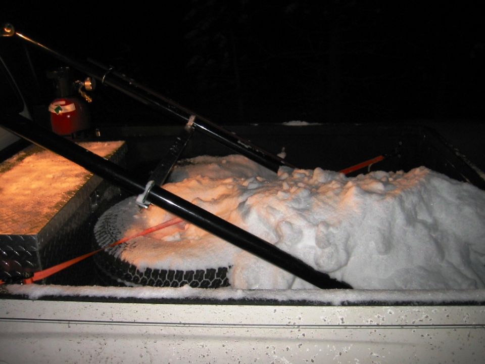 Snow-covered pickup bed at night with spare tire, ratchet strap, diamond-plate toolbox, and black rack bars