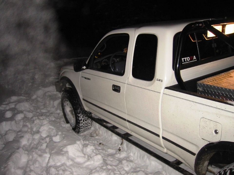 White Toyota Tacoma pickup truck in snow, side view. Features aftermarket wheels, tires with chains, and a roll bar