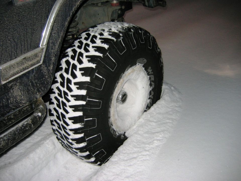 Truck tire with aggressive tread covered in snow, partially submerged in a snowdrift