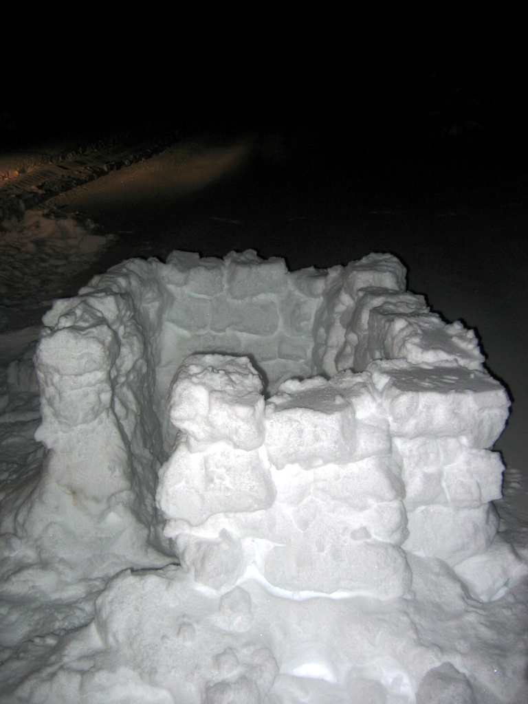 snow fort made of snow blocks at night, headlights visible in background