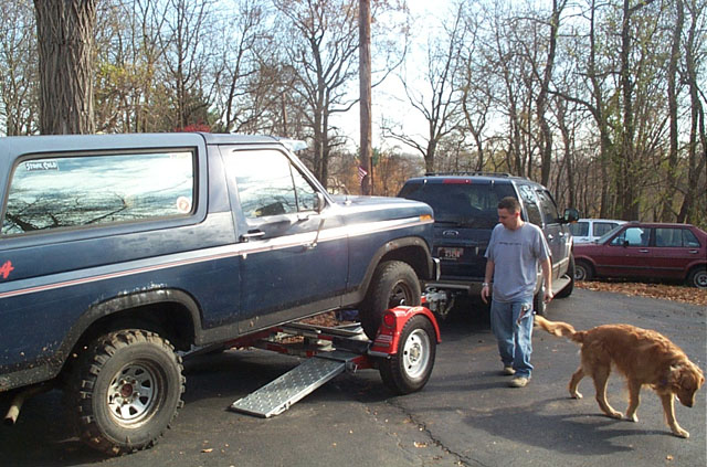 Blue Ford Bronco on a trailer, with a man and dog nearby. Other vehicles are parked in the background