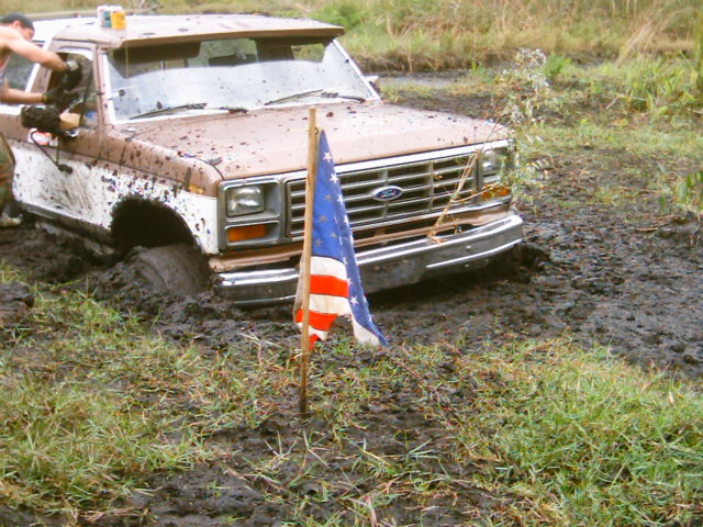 Mud-covered white Ford truck stuck axle-deep in a mud pit beside a small US flag on a stake