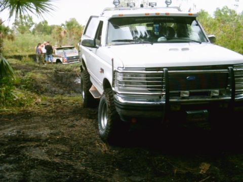 1995 Ford Bronco - Close front three-quarter view of a white lifted Ford Bronco parked in a muddy palmetto trail, showing its black tube brush guard, chrome b…