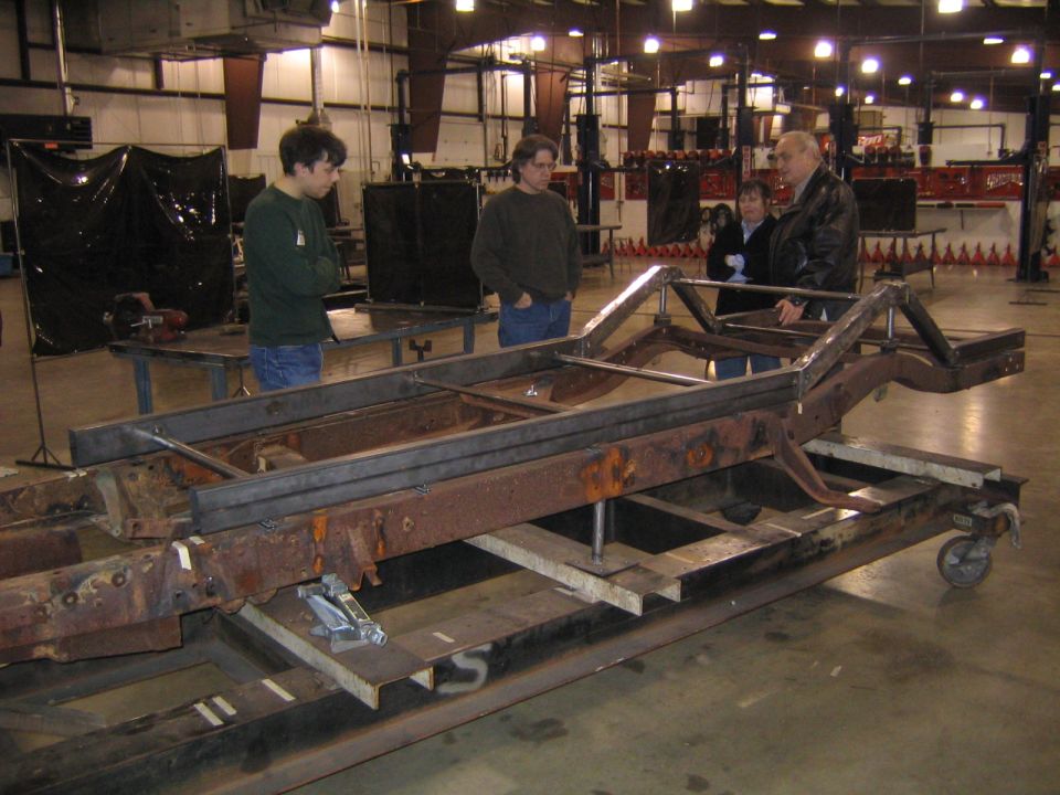 Rusty vehicle frame with metal bracing on a rolling cart, people observing