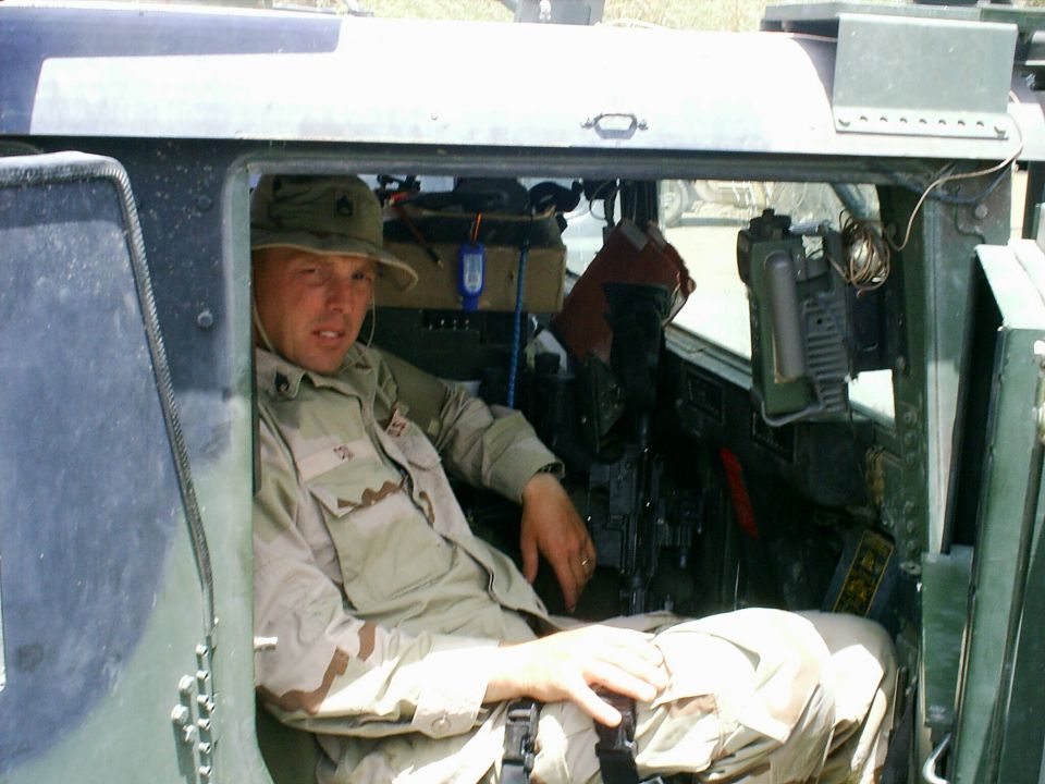 Soldier in desert camo seated inside a green military Humvee, seen through the open door.