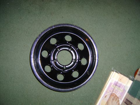 1985 Ford Bronco - A black wheel with chrome trim and ten circular cutouts is shown against a green background.