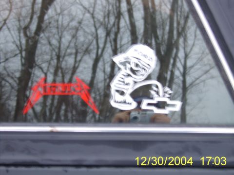 1985 Ford Bronco - Car window displays Metallica and Chevrolet stickers, with a reflection of trees and a camera.