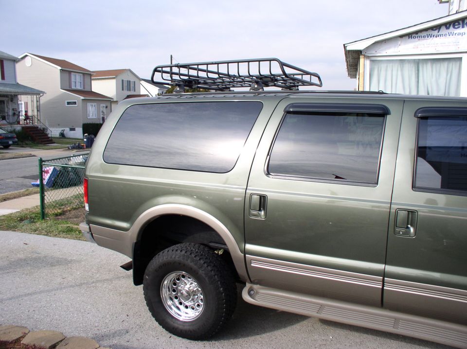 Green Ford Excursion SUV rear 3/4 view with roof basket and chrome wheels in a driveway.