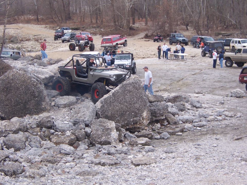 Gray doorless Jeep TJ with big off-road tires climbing boulders at a rocky off-road gathering