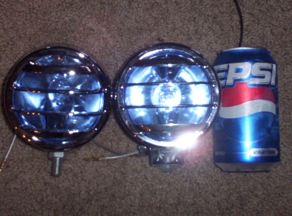 Two chrome-bezeled fog lights with blue lenses and protective grilles sit next to a Pepsi can