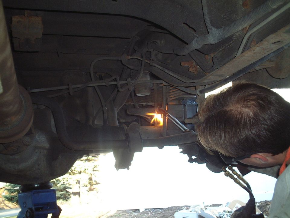 Man with torch heating rusty frame component under a raised vehicle