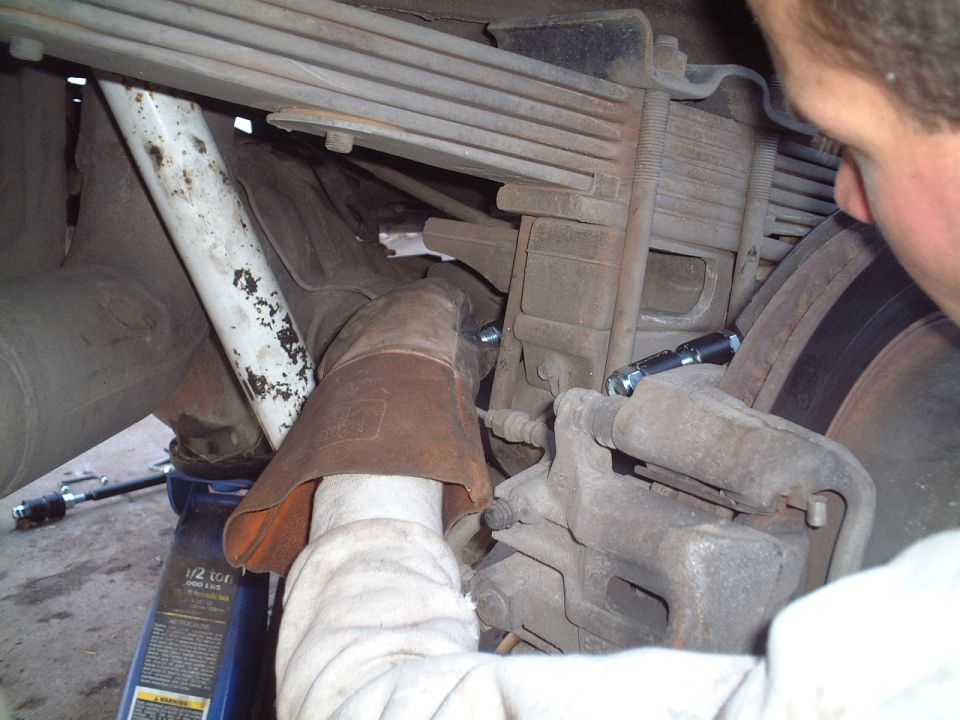 Underside view of rear brake caliper, leaf spring and axle with a blue bottle jack