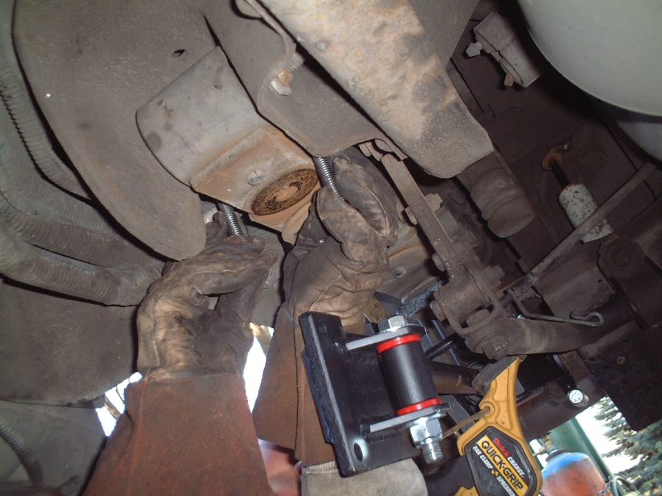 Gloved hands installing a black bushing mount under a vehicle, held by an orange Quick-Grip clamp.