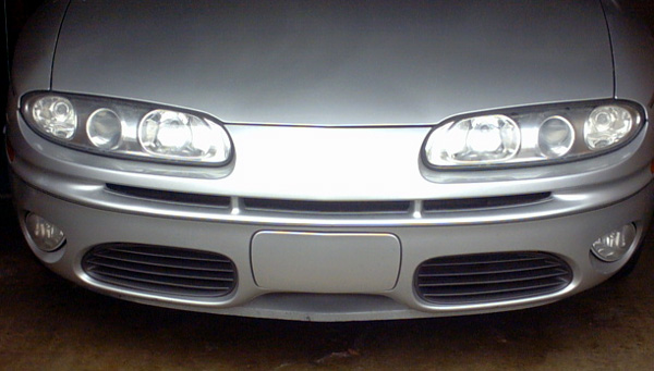 Front fascia of a silver sedan with quad headlights, lower grille openings, and fog lights in a garage