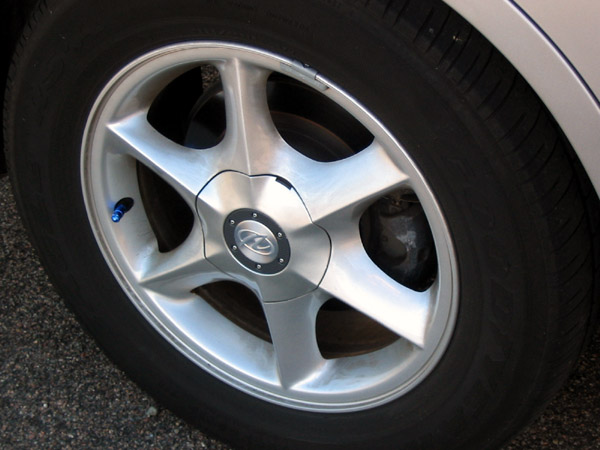 Six-spoke silver alloy wheel with Oldsmobile center cap and blue valve stem on a silver car