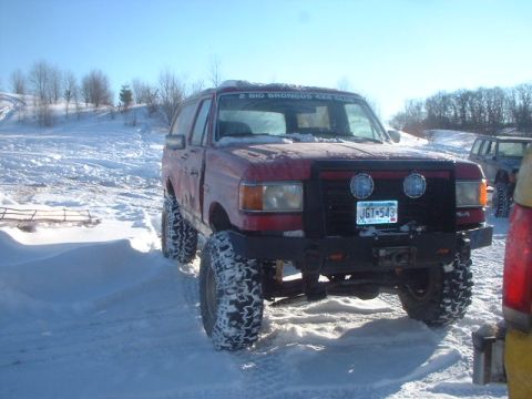 1989 Ford Bronco - A red SUV with large off-road tires sits in a snowy landscape, with another vehicle visible in the background.