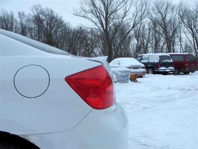 White sedan rear corner with taillight and fuel door in snowy lot, vans parked in background