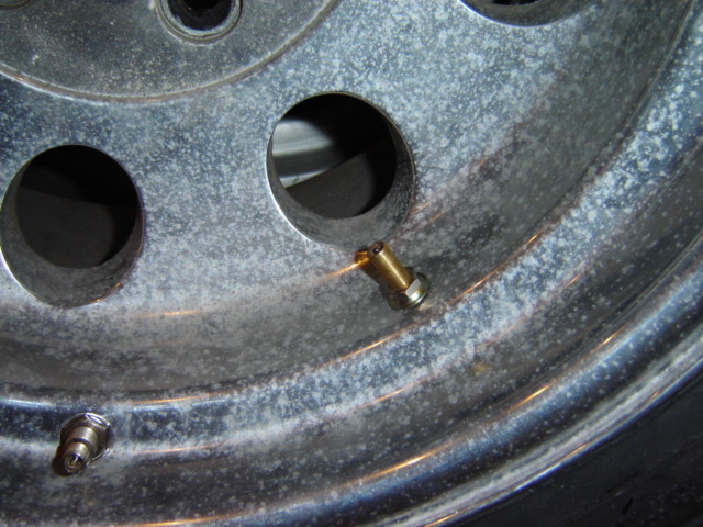Close-up of a corroded aluminum wheel showing two valve stems and lug nut holes. Supermotors.net logo in lower right