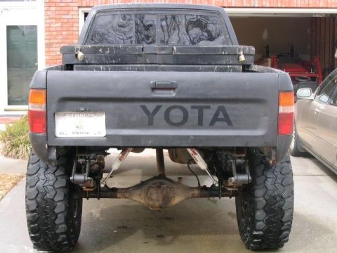 1986 Ford F-150 - Rear view of a black pickup truck with "YOTA" lettering and oversized tires, parked next to another vehicle.