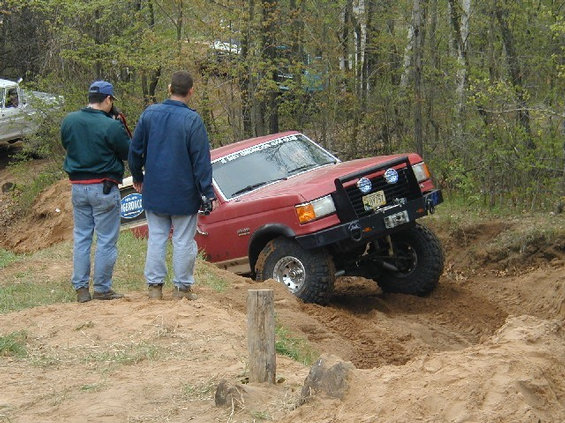 Red SUV stuck in dirt with two guys watching. Another white vehicle is in the background