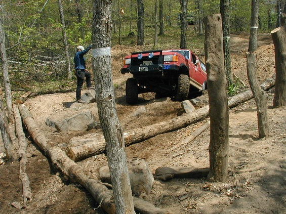 Red off-road vehicle with lifted suspension navigating a rocky, wooded trail