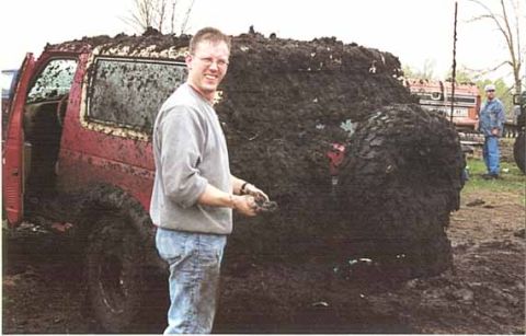 1989 Ford Bronco - A man stands beside a mud-covered 1989 Ford Bronco, its tires and body almost completely obscured by thick mud.