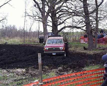 1989 Ford Bronco - A red 1989 Ford Bronco navigates a muddy off-road course, with trees and onlookers visible.