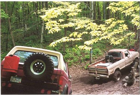 1989 Ford Bronco - Two off-road vehicles navigate a muddy path through a wooded area.