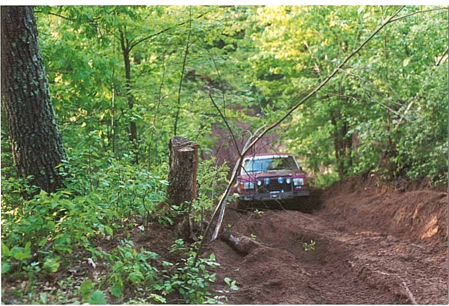 Jeep Cherokee XJ driving up a muddy trail in a forest. Watermark reads SUPERMOTORS