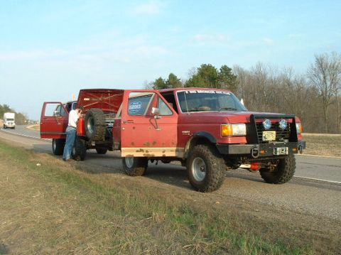 1989 Ford Bronco - A red 1989 Ford Bronco is stopped on the roadside, towing a trailer with its rear hatch open.