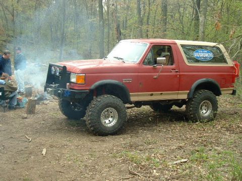1989 Ford Bronco - A red Ford Bronco is parked in a wooded area with people gathered around a campfire.