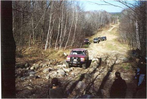 1989 Ford Bronco - A convoy of SUVs navigates a challenging off-road trail in a wooded area.
