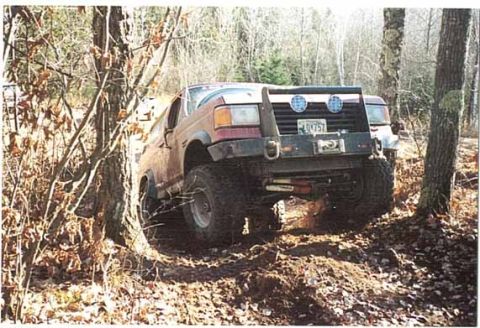 1989 Ford Bronco - A red Ford Bronco is shown off-roading on a dirt path surrounded by trees.