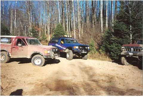 1989 Ford Bronco - Multiple off-road SUVs are parked on a dirt path surrounded by trees.