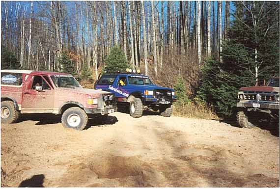 Two off-road SUVs, one red and one blue, parked on a dirt path with trees in the background. The red SUV appears to be a 1987 Ford Bronco