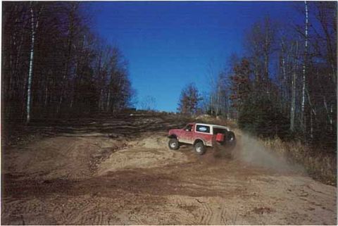 1989 Ford Bronco - A red SUV navigates a dusty off-road trail under a clear blue sky.