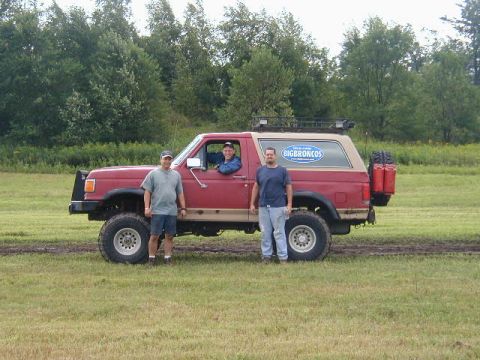 1989 Ford Bronco - A red Ford Bronco with a tan top is parked on a muddy field with three men standing around it.