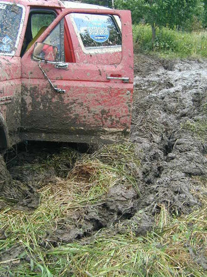 1989 Ford Bronco - Muddy red Ford F-Series truck stuck in a ditch with grass and thick mud.