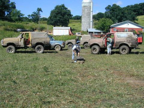 1989 Ford Bronco - Several muddy off-road vehicles, including a 1987 Ford Bronco, are parked in a grassy field near a barn and silo.