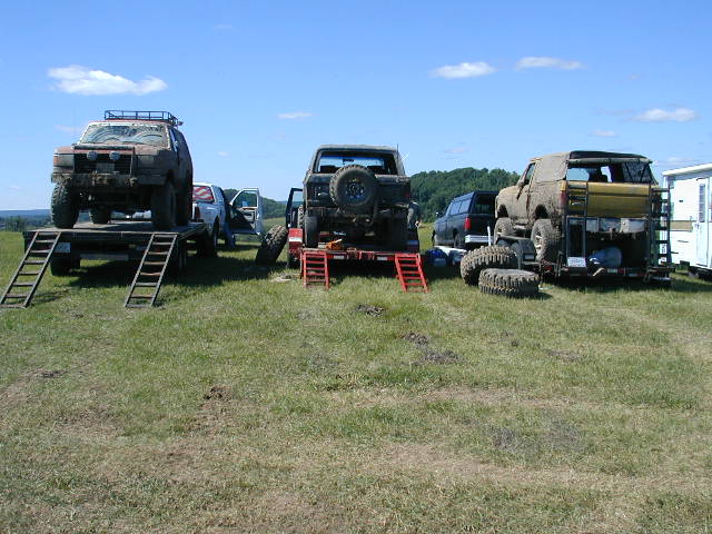 Three off-road vehicles are loaded onto trailers in a grassy field under a blue sky