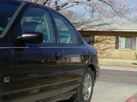1995 Ford Contour - Side view of a dark sedan, with a house in the background. The registry claims it is a 1995 Ford Contour, but this is not confirmed by the…