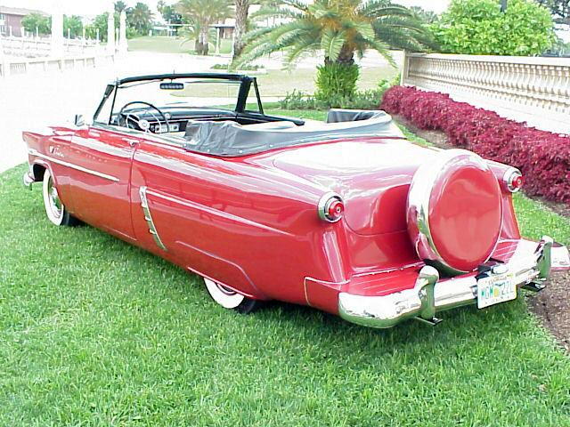 Red 1950s Ford convertible, top down, rear 3/4 view with continental kit and whitewall tires on grass