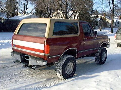 1991 Ford Bronco - A red and tan 1991 Ford Bronco equipped with large tires sits in a snowy landscape.